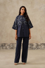 A woman modeling a navy blue two-piece set consisting of a short-sleeved button-up top with a white floral pattern and matching navy wide-leg trousers with lace trim, set against a textured beige studio backdrop.