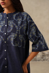 A close-up of a woman wearing a navy blue button-front tunic or kurta with a light blue and sage paisley embroidery, cropped from the lower face to the hips against a neutral textured wall.