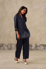 A woman modeling a navy blue two-piece outfit consisting of a long-sleeve tunic top with floral embroidery and matching wide-leg trousers, set against a textured beige studio backdrop. denim coord set, cutwork embroidery