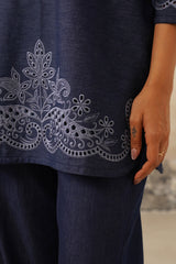 Close-up of a navy blue tunic or kurta with intricate white floral embroidery and eyelet cutouts along the hem, worn with matching navy pants. A person's hand is resting near the hem.