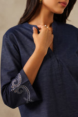 Close-up of a woman wearing a navy blue tunic top with tonal embroidered detailing on the sleeve, set against a neutral beige background.