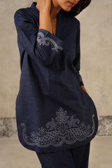 A woman wearing a dark navy denim blue tunic top with intricate silver-gray embroidery on the sleeve and hem, paired with matching navy pants, set against a textured beige wall.