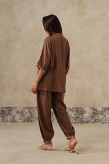 A woman shown from the back wearing a matching brown two-piece lounge set consisting of a loose short-sleeve top and wide-leg pants, paired with beige heeled sandals against a textured beige wall.