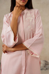 A close-up of a woman wearing a light pink traditional tunic with white floral embroidery on the chest and scalloped white trim on the wide sleeves, set against a neutral textured background.