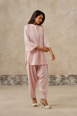 A woman modeling a light pink traditional outfit with matching pants and white heeled sandals, standing against a textured beige wall.