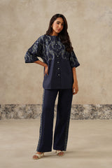 A woman modeling a denim coord set consisting of a short-sleeved button-up top with white paisley embroidery and matching wide-leg trousers with embroidered side details, set against a textured beige studio backdrop.