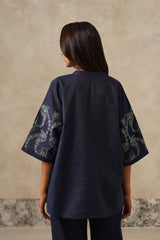 A woman viewed from the back wearing a dark navy blue short-sleeve top with ornate light-colored paisley embroidery on the sleeves, set against a textured beige wall.
