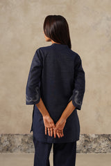 A woman viewed from behind wearing a dark navy blue kurta with three-quarter sleeves and white embroidered details on the cuffs, paired with matching navy trousers, set against a textured beige wall.