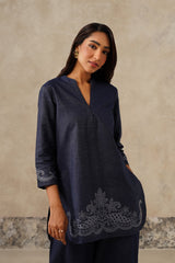 A woman modeling a navy blue denim tunic top with tonal cutwork embroidered detailing on the cuffs and hem, set against a textured beige wall.