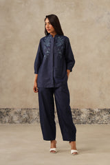 A woman modeling a denim coord set consisting of a long-sleeve button-up top with floral embroidery on the chest and matching wide-leg cropped pants, set against a textured beige studio backdrop.