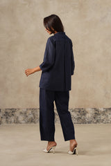 A woman is shown from the back wearing a dark navy blue two-piece set consisting of a long-sleeve button-up shirt and matching wide-leg trousers, paired with white high-heeled sandals, set against a textured beige studio backdrop.
