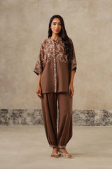 A woman modeling a brown linen coord set consisting of a short-sleeved button-up kurta with intricate embroidery and matching wide-leg harems, set against a textured beige studio backdrop.