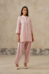 A woman modeling a light pink line coord set outfit consisting of a long-sleeve embroidered kurta and matching wide-leg pants, standing against a textured beige studio wall.